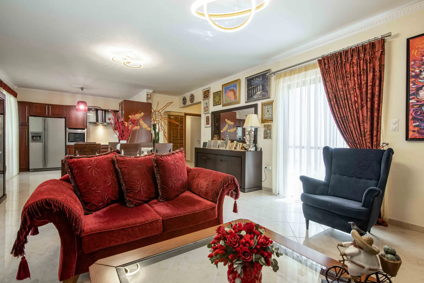 Living room with a red velvet sofa, dark armchair, coffee table with red flowers, and an open kitchen in the background.