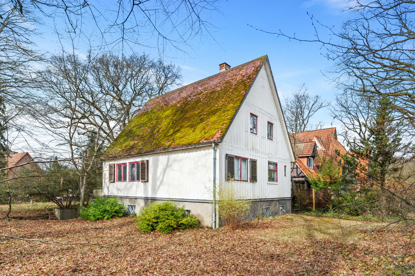 Exterior of a white house with a mossy roof, red window frames, and brown shutters.