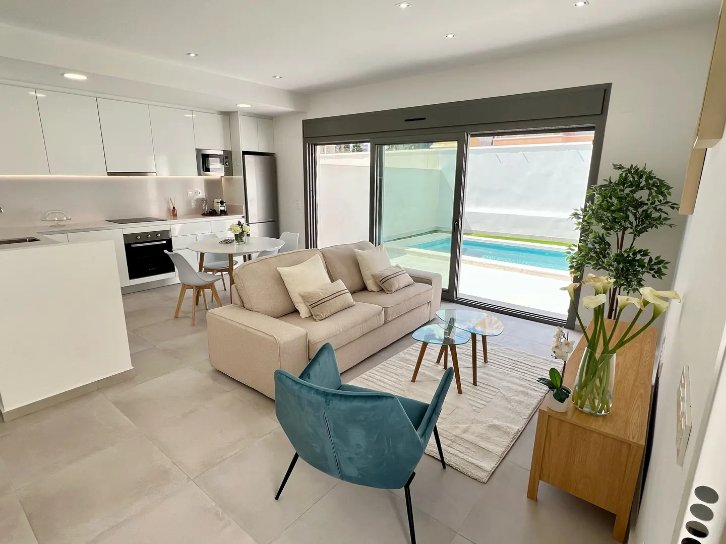 Bright, modern living space with a beige sofa, blue chair, and glass tables. Kitchen with white cabinets and a view of a pool through sliding glass doors.