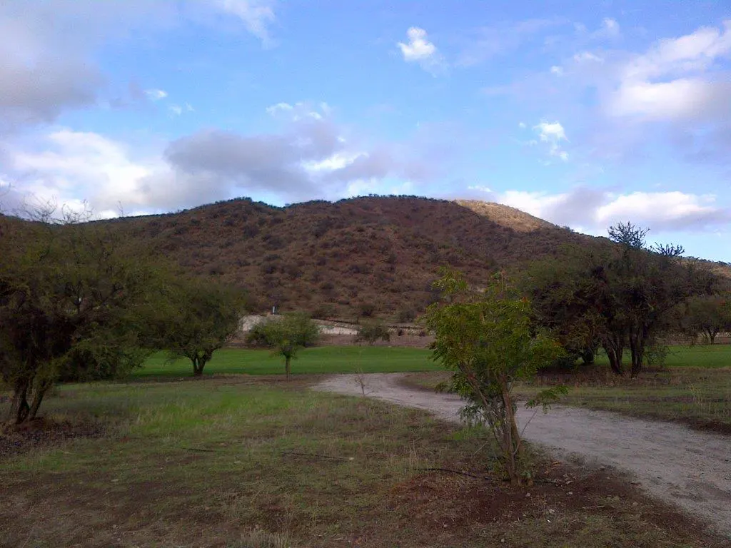 Landscape view of a brown mountain under a blue sky with clouds, green grass, trees, and a dirt path.