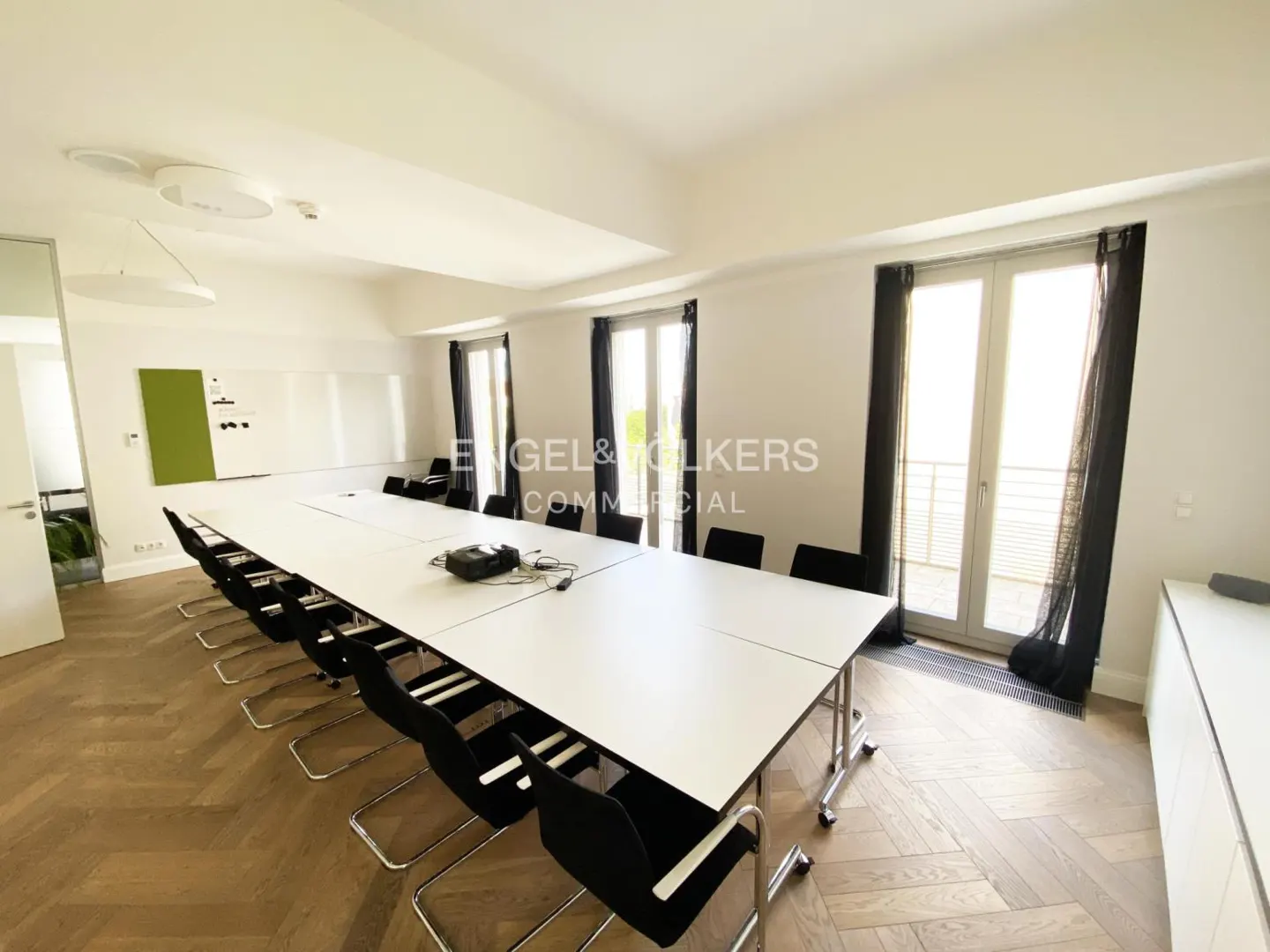 Bright conference room with a long white table, black chairs, herringbone wood floor, and white walls. Windows with dark curtains.