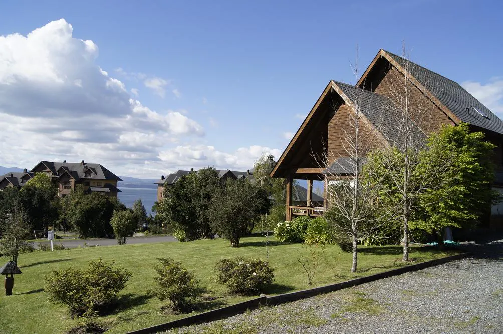 A brown wooden house with a dark roof sits on a green lawn under a blue sky with white clouds. Other houses and a lake are in the background.