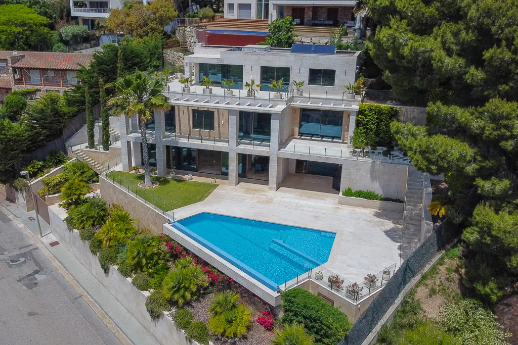 Aerial view of a modern, multi-story beige house with a blue swimming pool, surrounded by lush greenery and other houses.