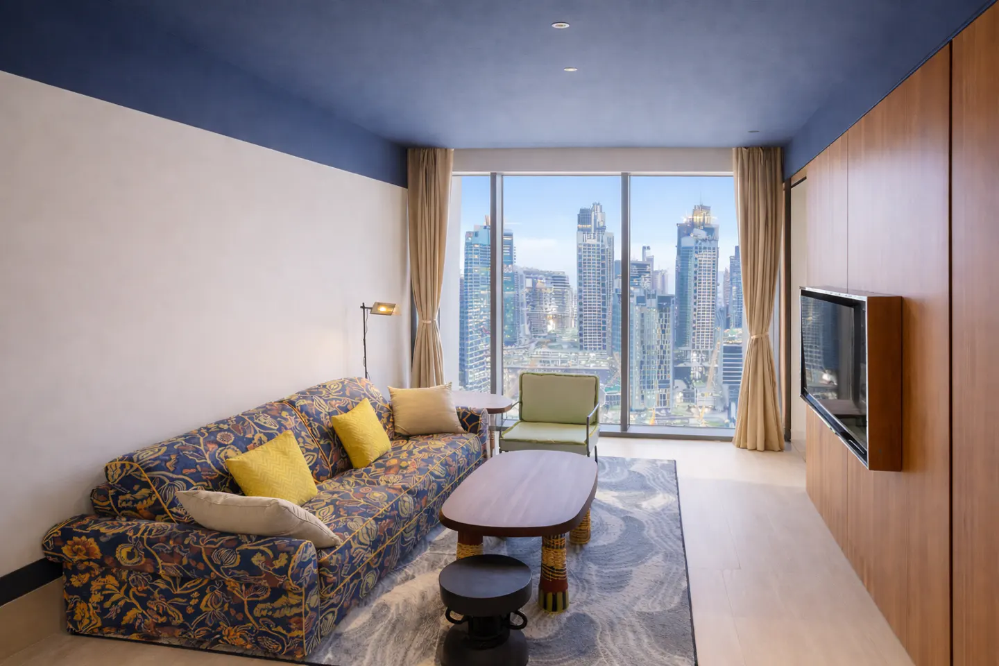 Living room with a floral sofa, yellow pillows, and a view of city skyscrapers through a large window.
