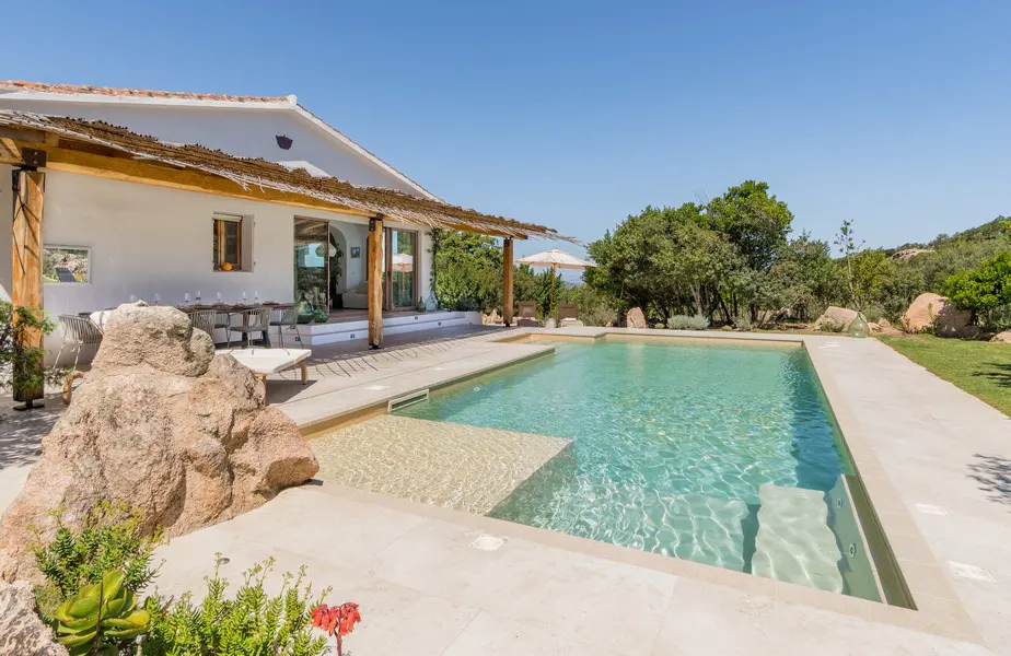 A white villa with a pool on a sunny day. The pool has a shallow end and is surrounded by stone. A dining table is on the patio.