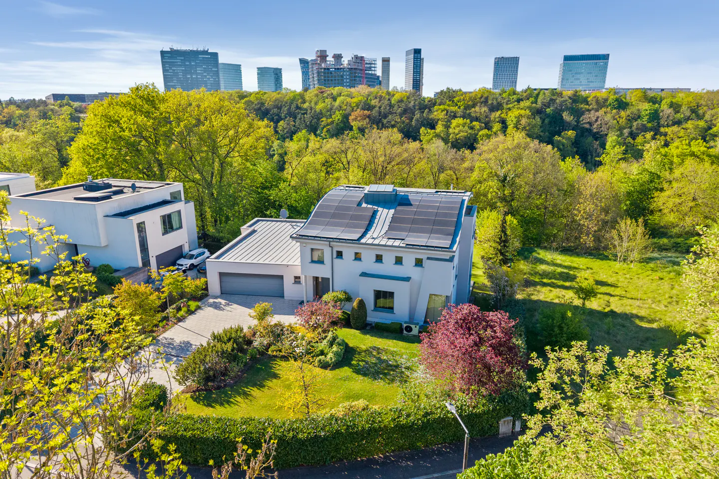 Aerial view of a modern white house with solar panels, a garage, and lush green landscaping. Tall buildings are visible in the background.