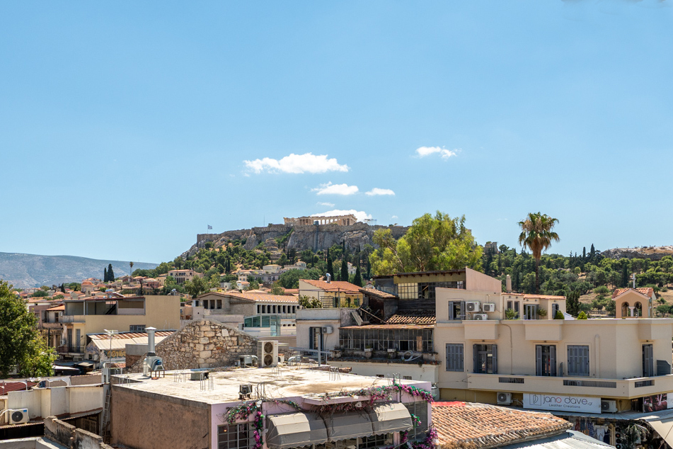 View of the Acropolis in Athens rising above a dense urban neighborhood with tan buildings and a clear blue sky.