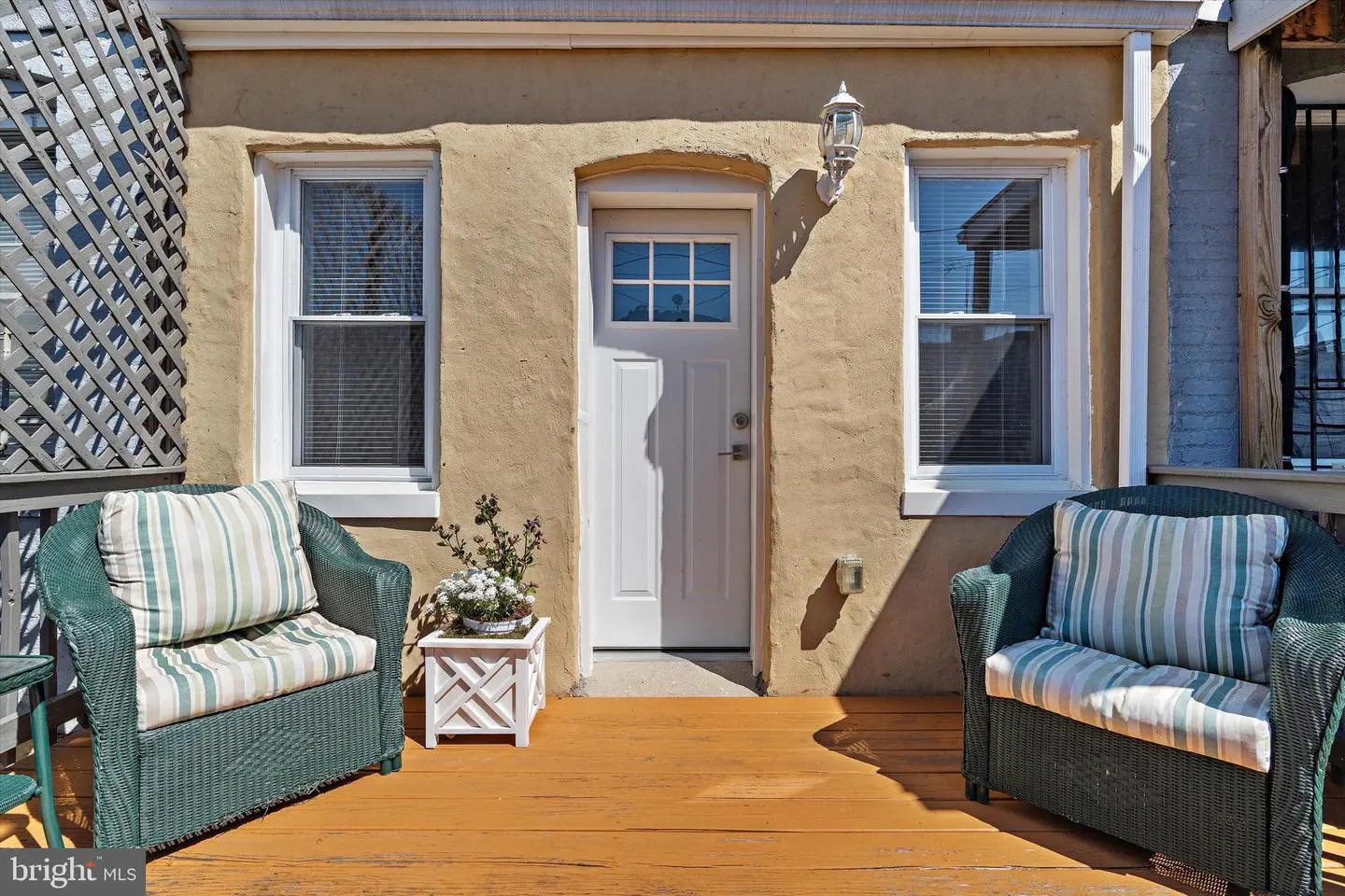 Outdoor patio with two green wicker chairs, striped cushions, and a white flower box. A white door is centered between two windows.