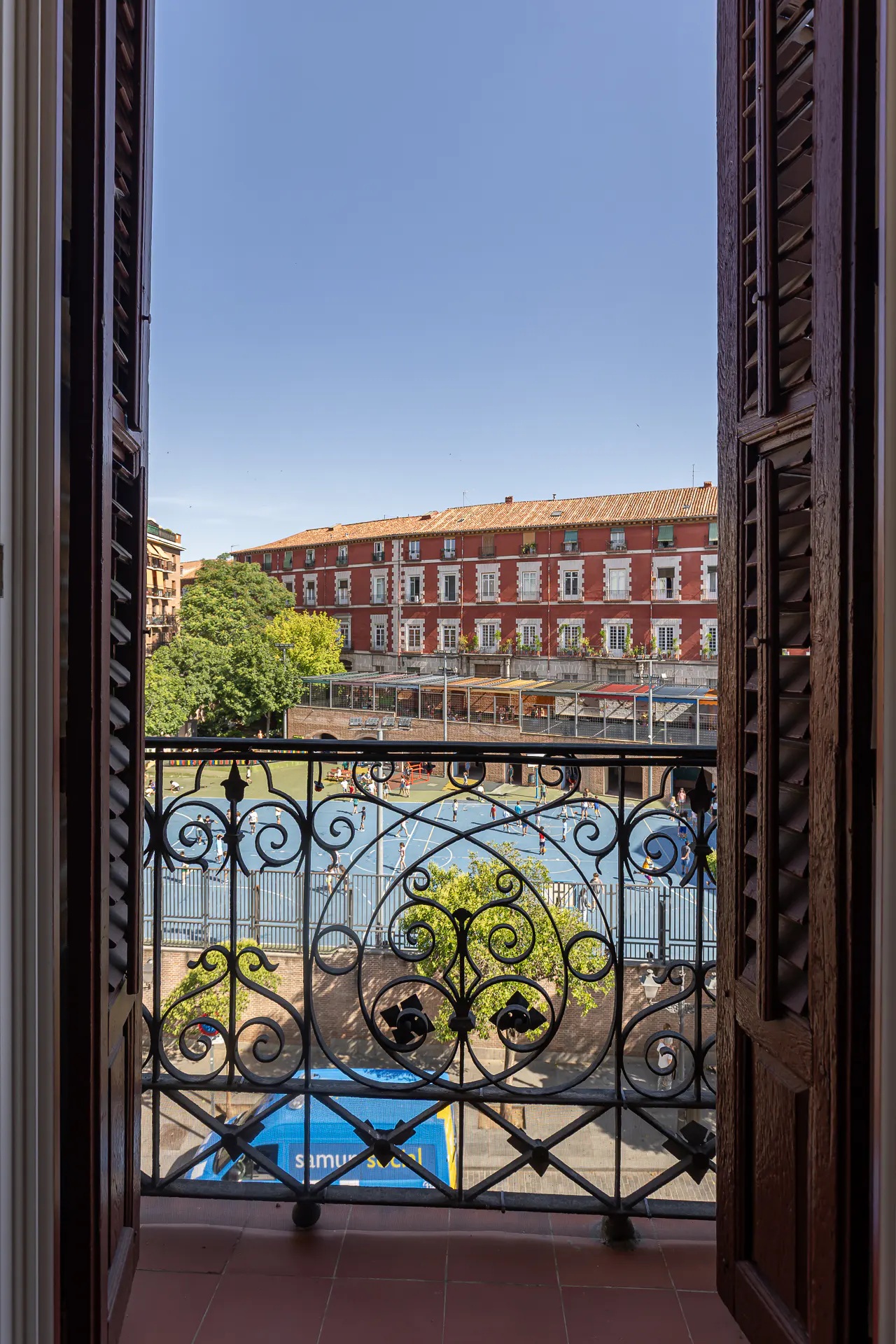 View from a balcony with open brown shutters, overlooking a blue sports field, trees, and a red brick building under a clear blue sky.