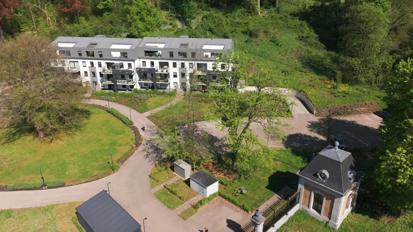 Aerial view of modern white apartments with gray roofs, surrounded by green lawns, trees, and a small brick building.