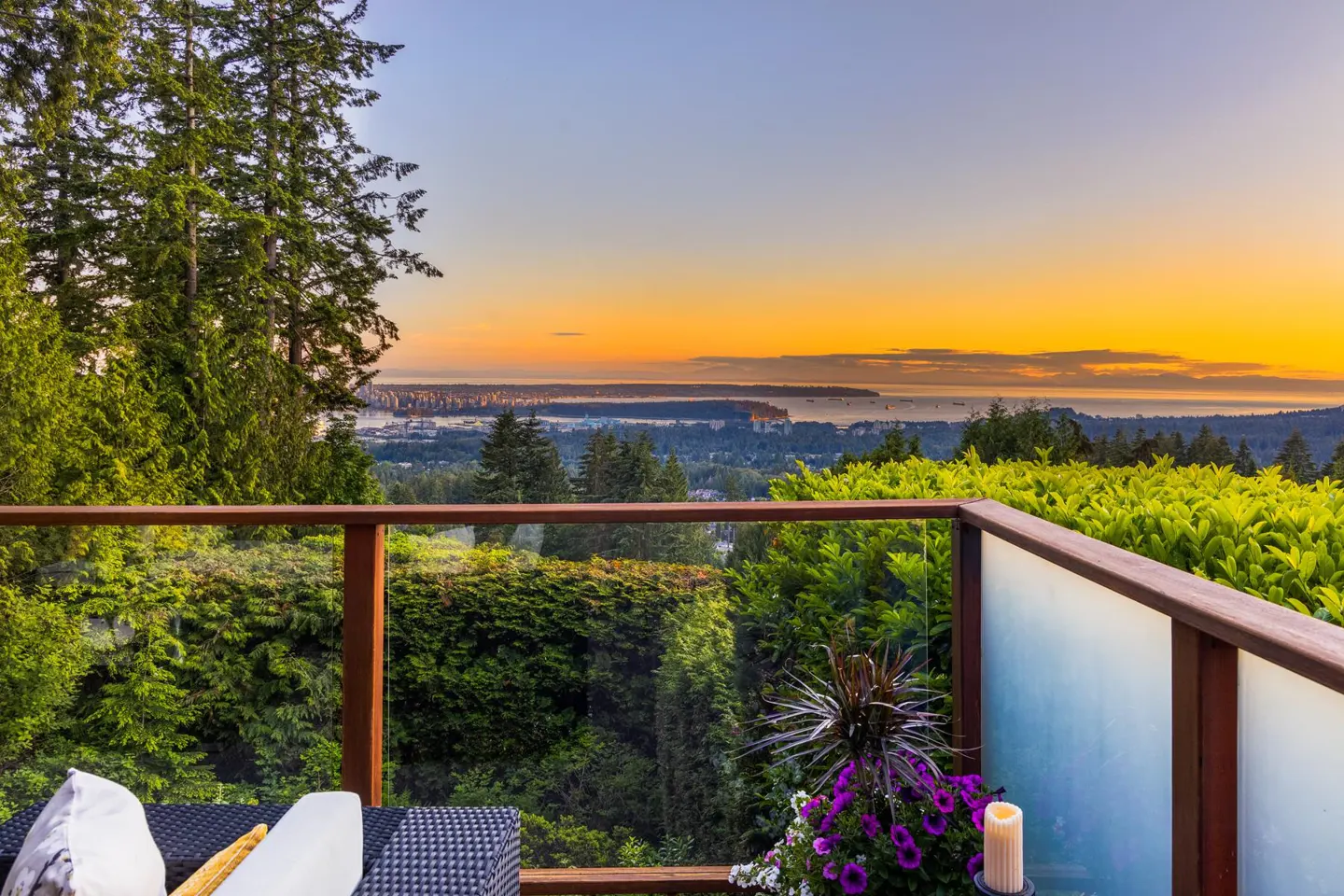 Balcony view at sunset. Glass railing, wicker furniture, and purple flowers overlook a city skyline and ocean.