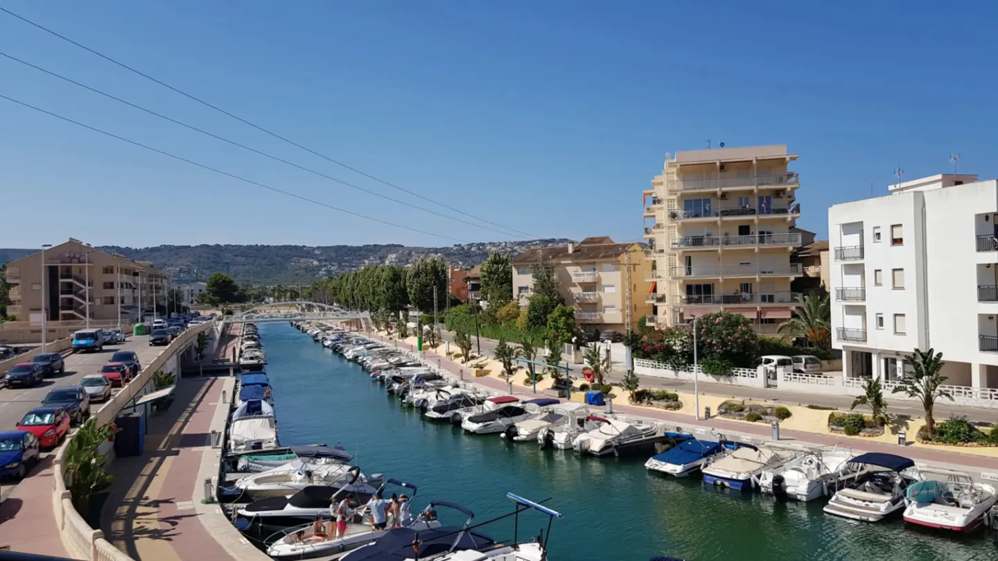 A canal lined with docked boats, flanked by apartment buildings, a road with cars, and a clear blue sky.