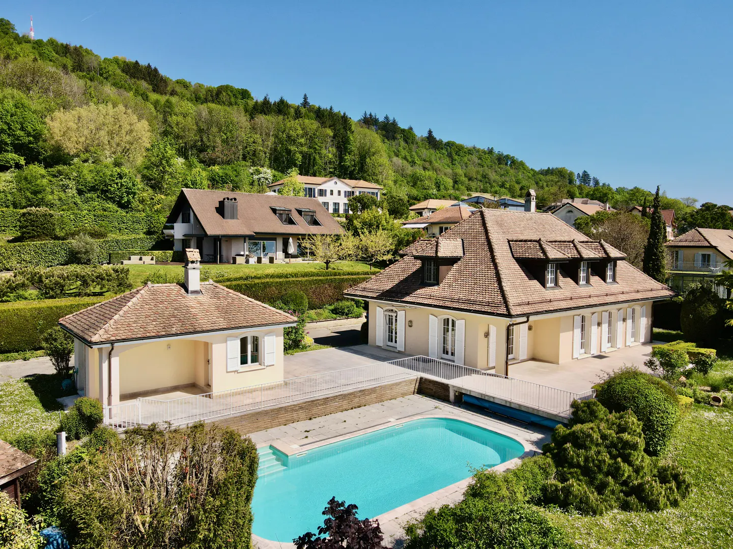 Aerial view of a beige house with a brown roof and a blue pool, surrounded by green trees and a hill.