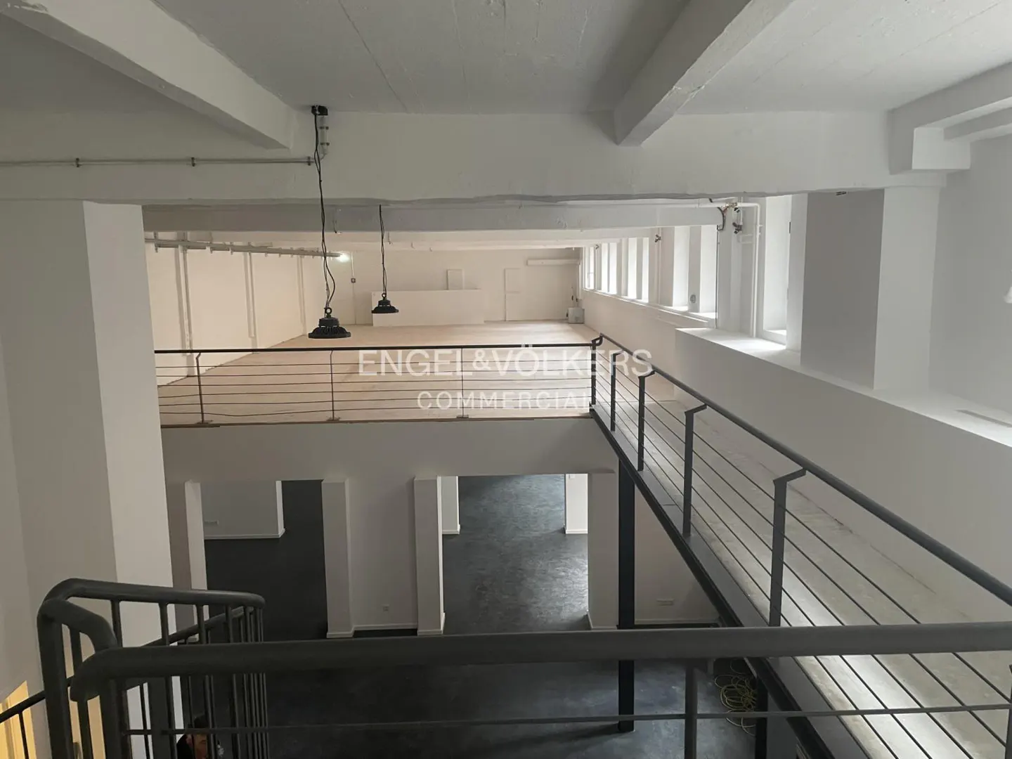 Interior view of a commercial space with white walls, dark floors, and a mezzanine level with metal railings. Stairs lead down to the lower level.