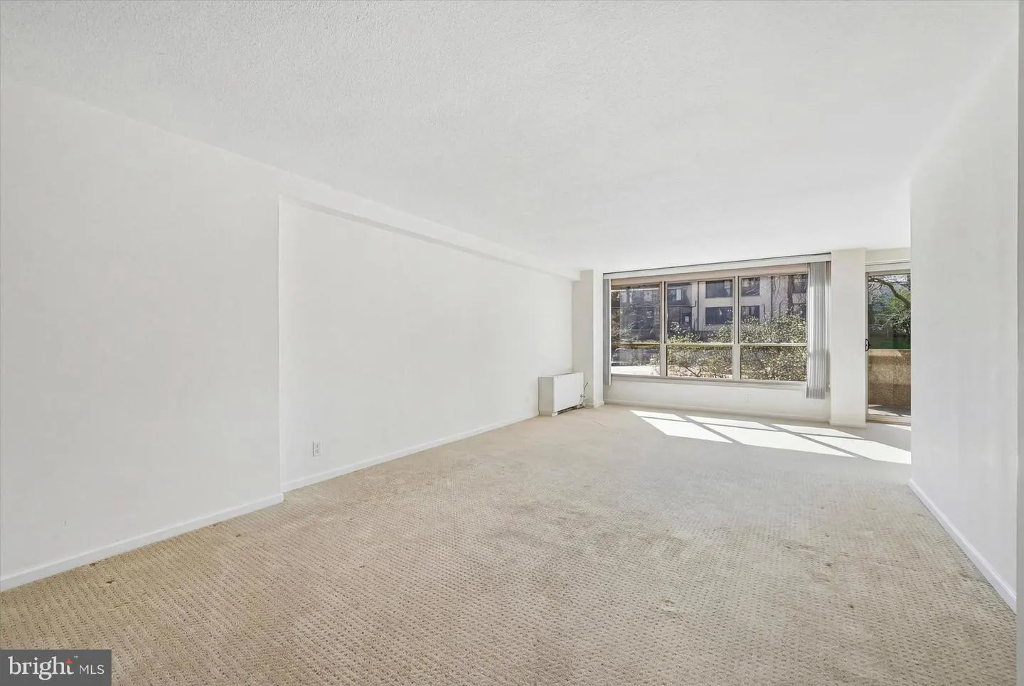 Bright, empty room with beige carpet, white walls, and large windows showing an apartment building and trees.