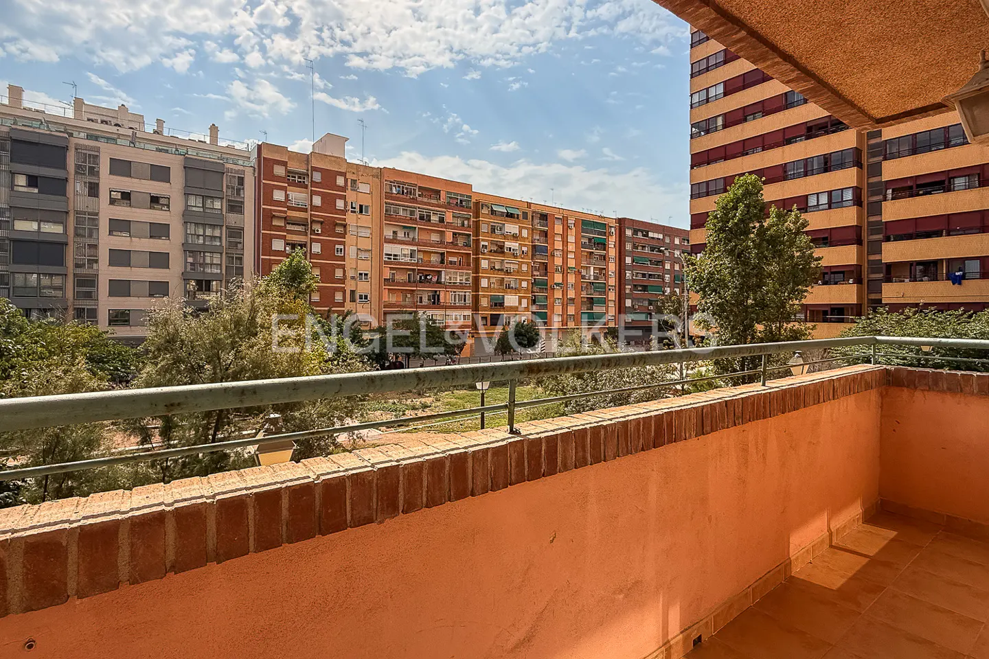 View from a balcony with a brick railing, overlooking a park and apartment buildings under a blue sky.