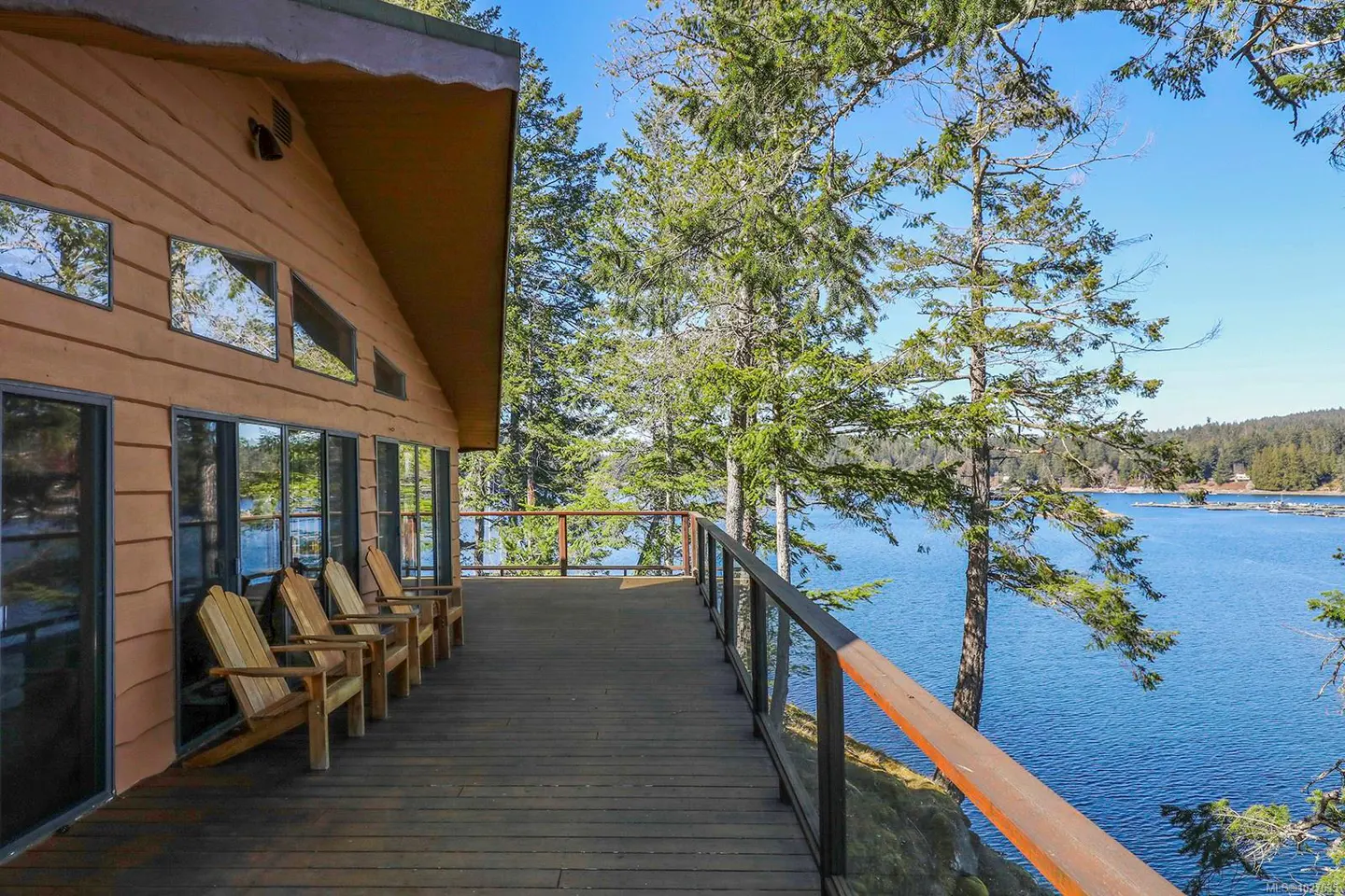 Exterior view of a wood cabin with a deck overlooking a blue lake and green trees on a sunny day. Four wooden chairs are on the deck.