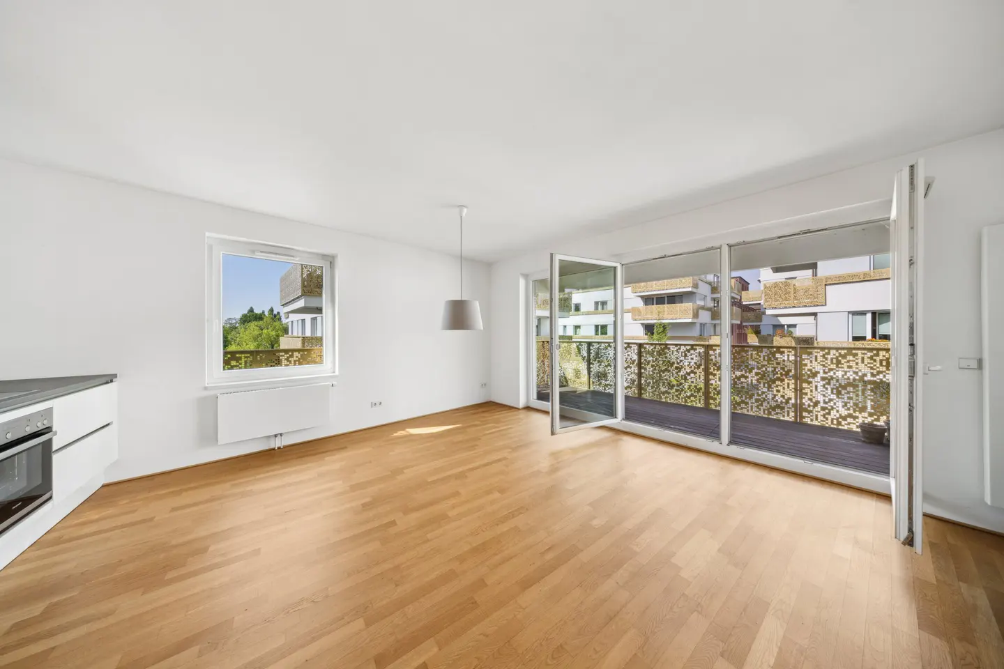 Bright, empty apartment with wood floors, white walls, and a balcony with decorative railings. A window and kitchen appliances are visible.