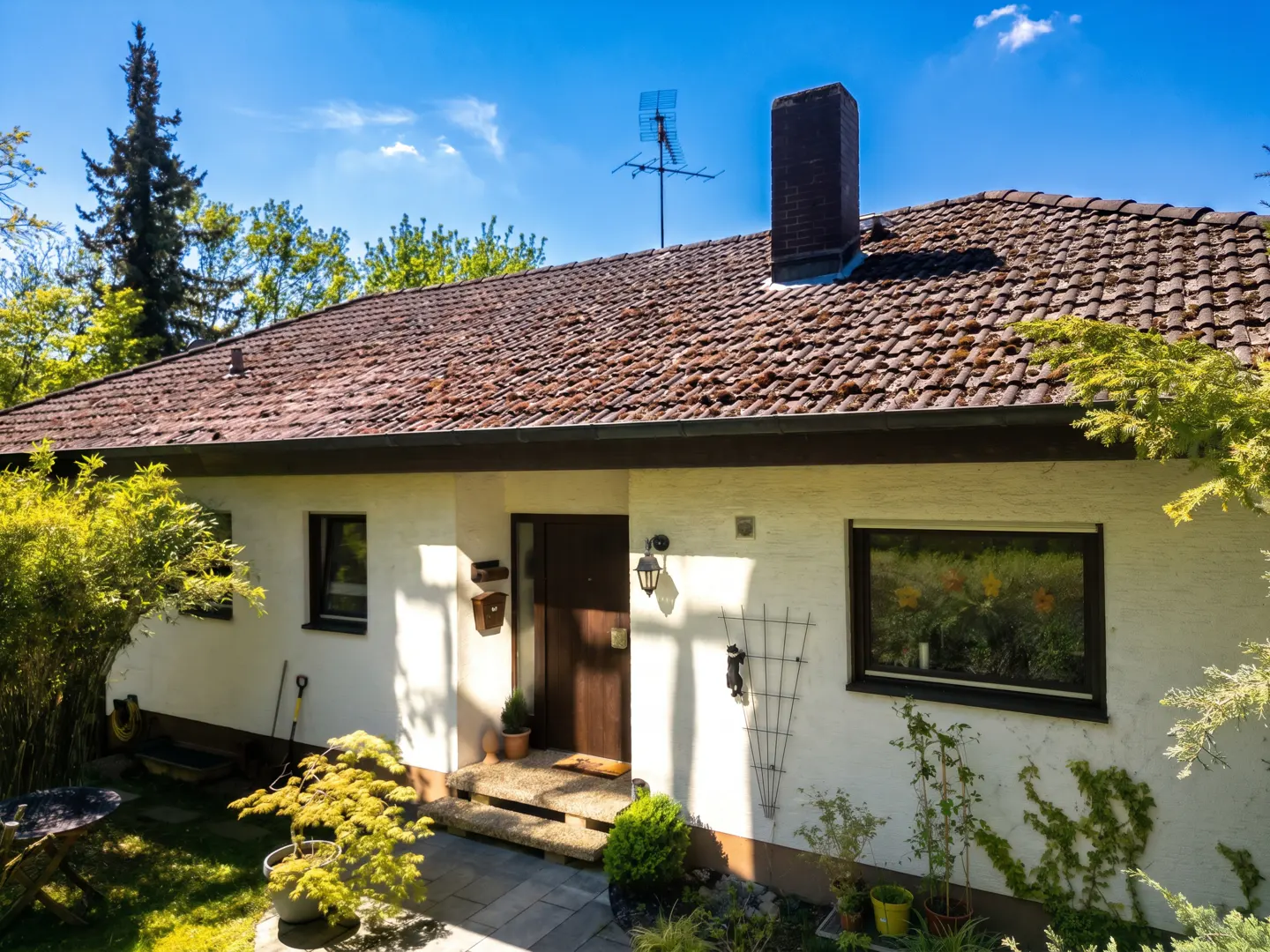 A single-story white house with a brown tile roof, chimney, and antenna under a blue sky. The house has dark-framed windows and a brown front door. Green trees surround the house.