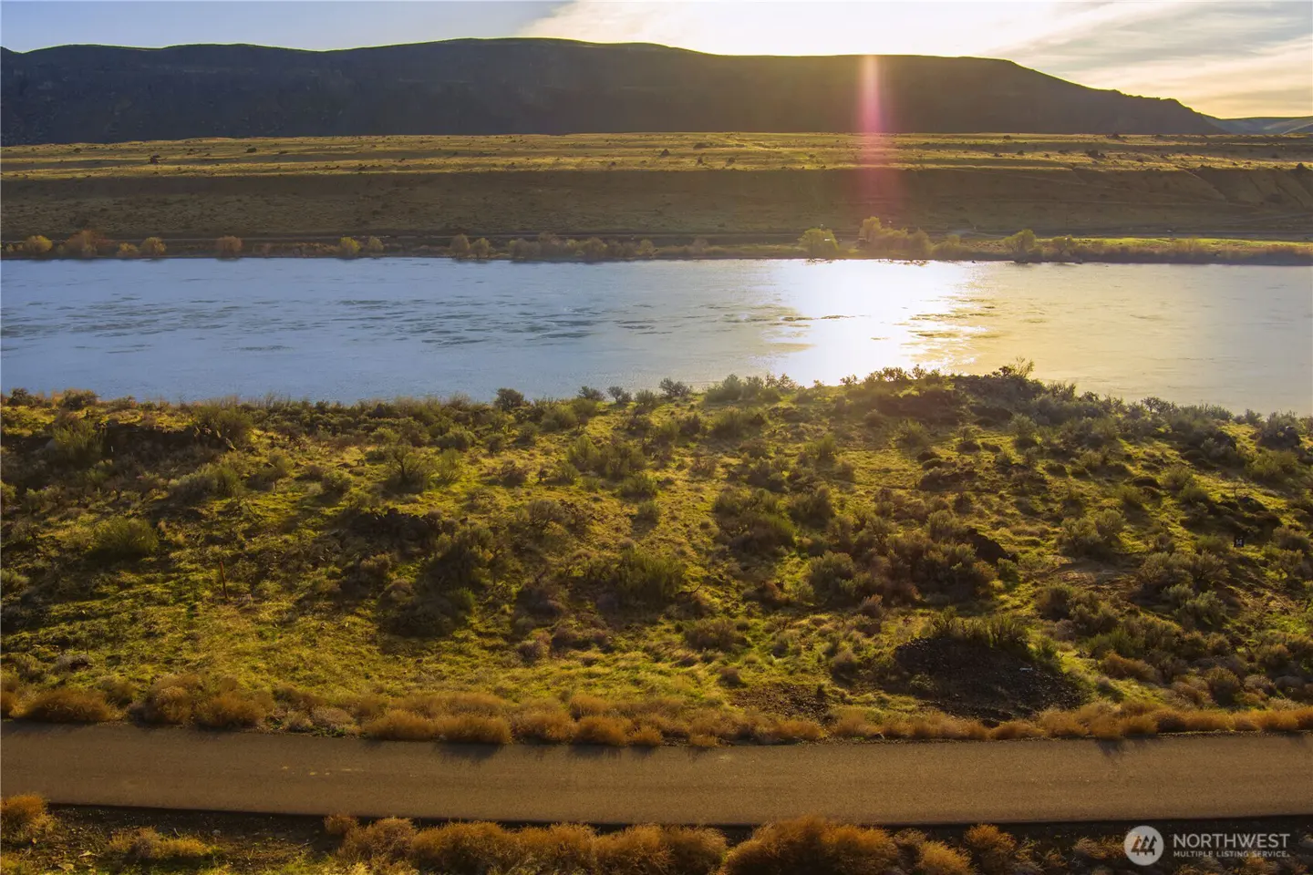 Scenic view of a wide river reflecting sunlight, with green hills and a road in the foreground.