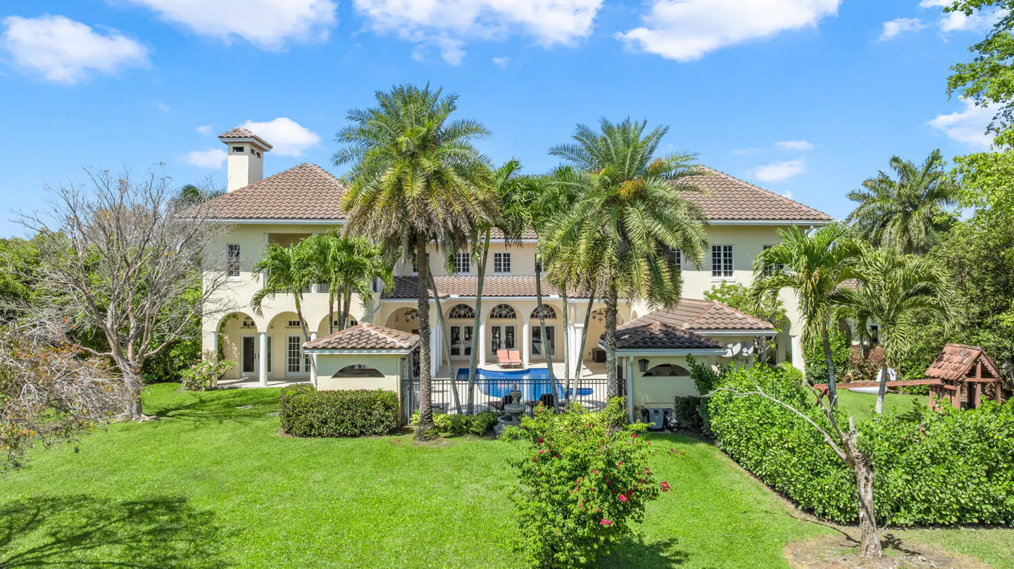 Rear view of a large, light yellow house with a brown tile roof, palm trees, a pool, and green lawn under a blue sky.
