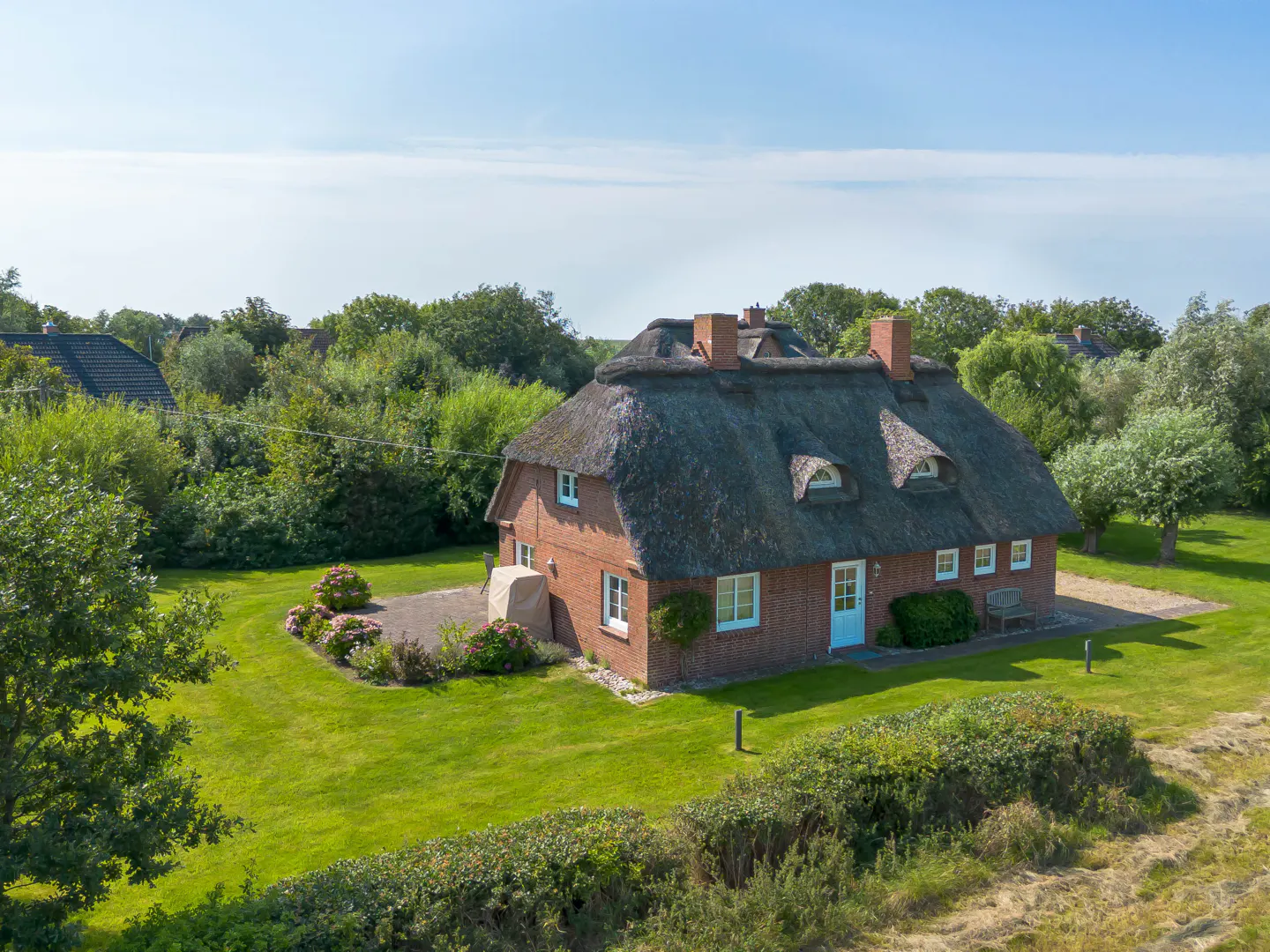 A red brick house with a thatched roof sits on a green lawn surrounded by trees under a blue sky.
