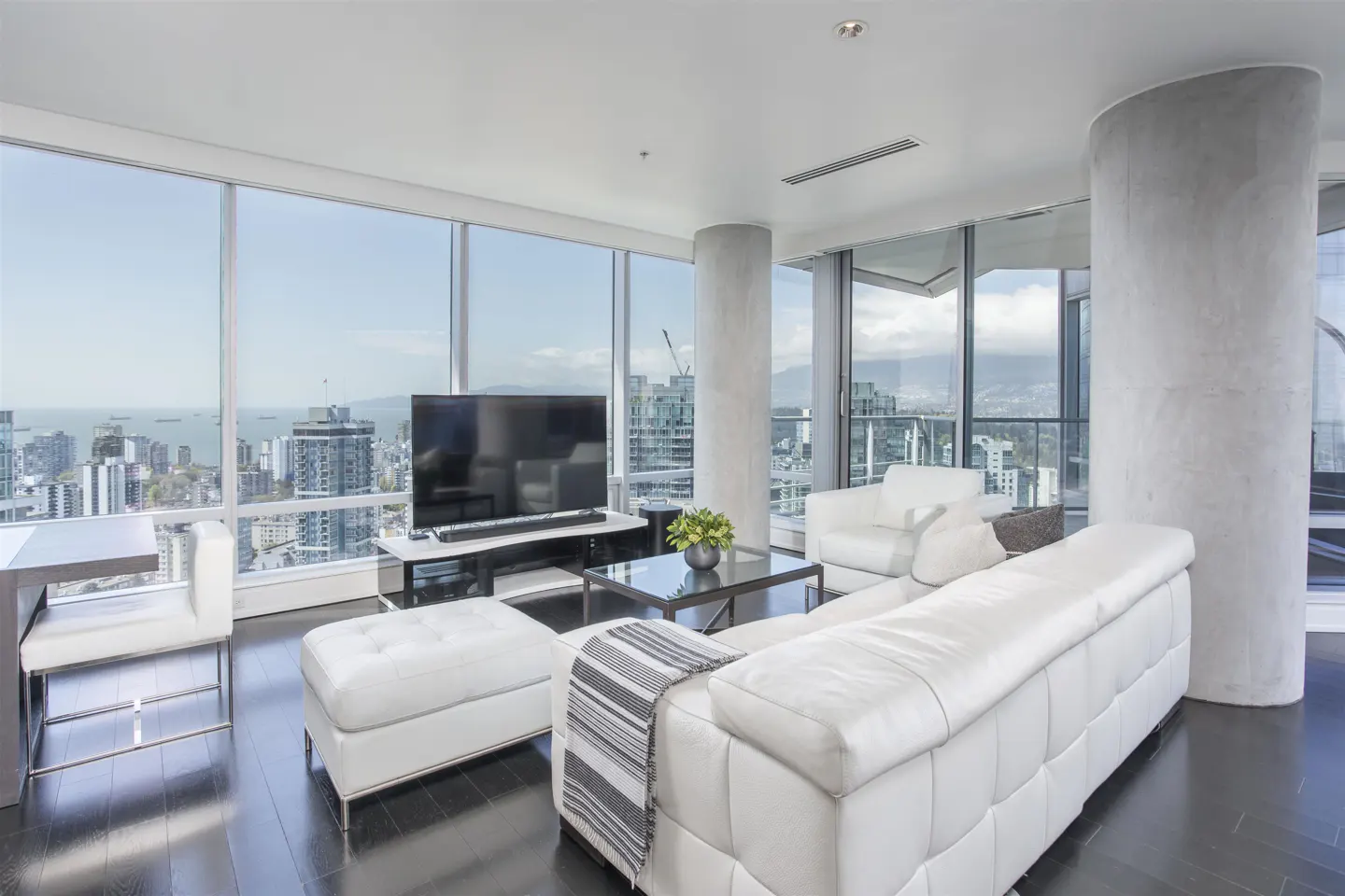 Bright living room with floor-to-ceiling windows showing a city view. White sofa, chair, and ottoman on dark wood floors.