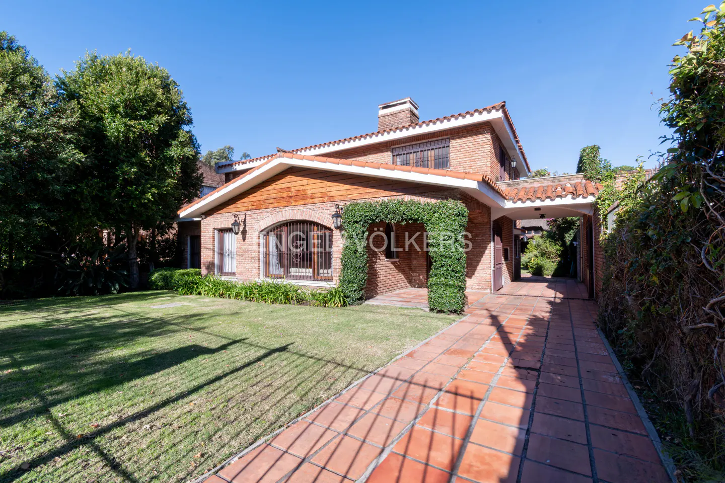 Two-story brick house with a red tile roof, green lawn, and brick walkway on a sunny day. The entrance is framed by greenery.