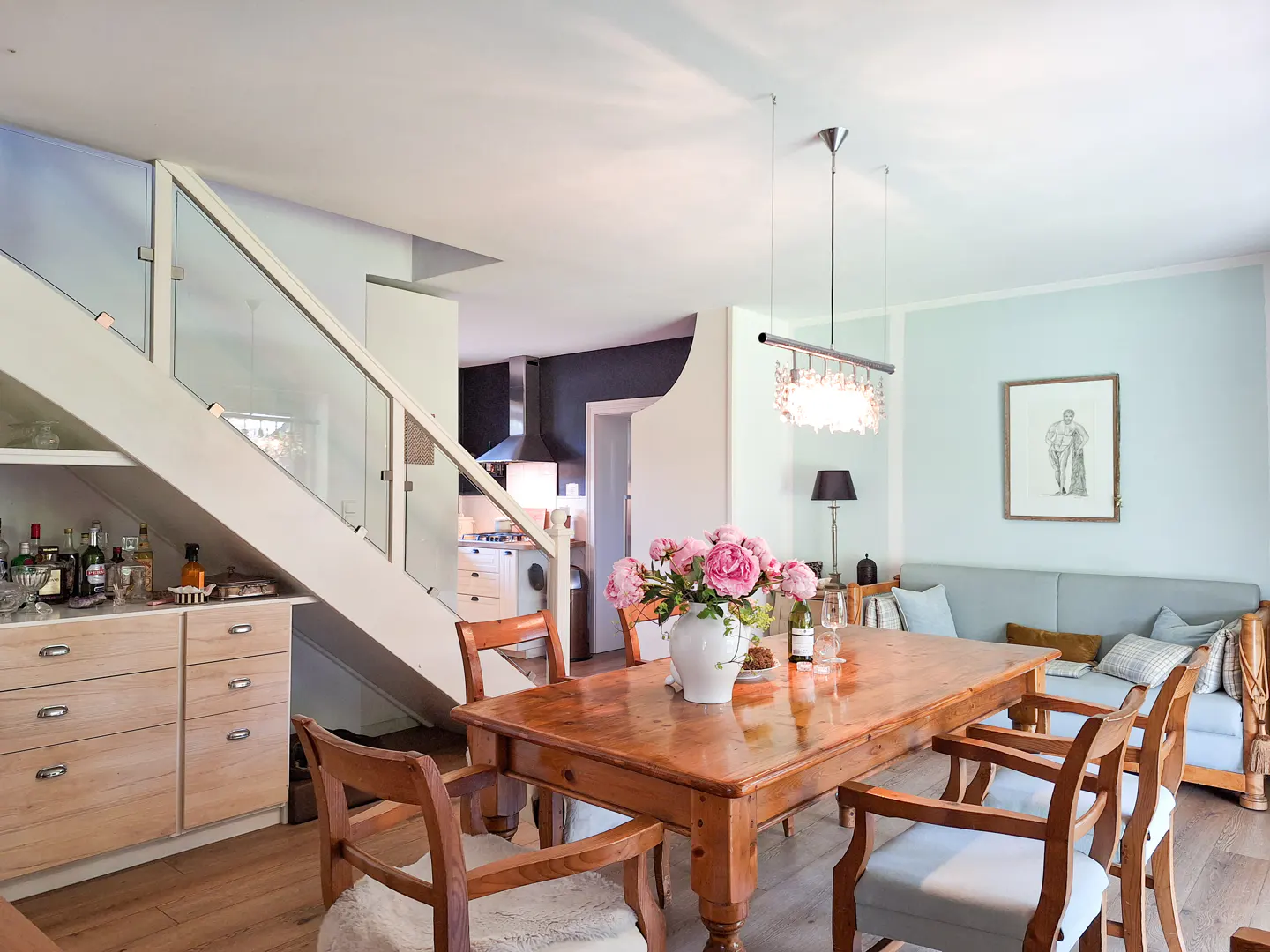 Open-concept dining area with a wooden table, chairs, and a vase of pink roses. Stairs with glass railings lead to the upper level. Light blue walls and wooden floors.