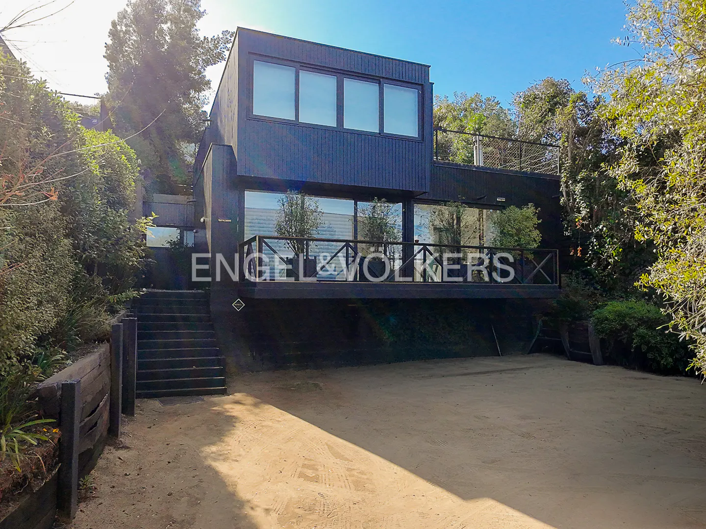 Modern black house with large windows and a deck, surrounded by trees and greenery. Stairs lead up to the house. The ground is sandy.