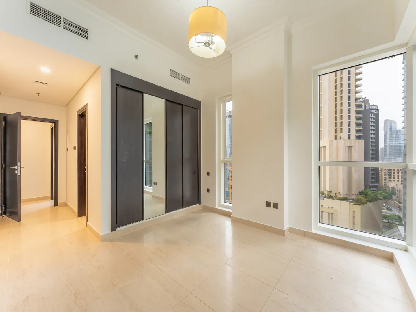 A bright, empty room with beige tile floors, a large window with a city view, and a dark sliding-door closet.