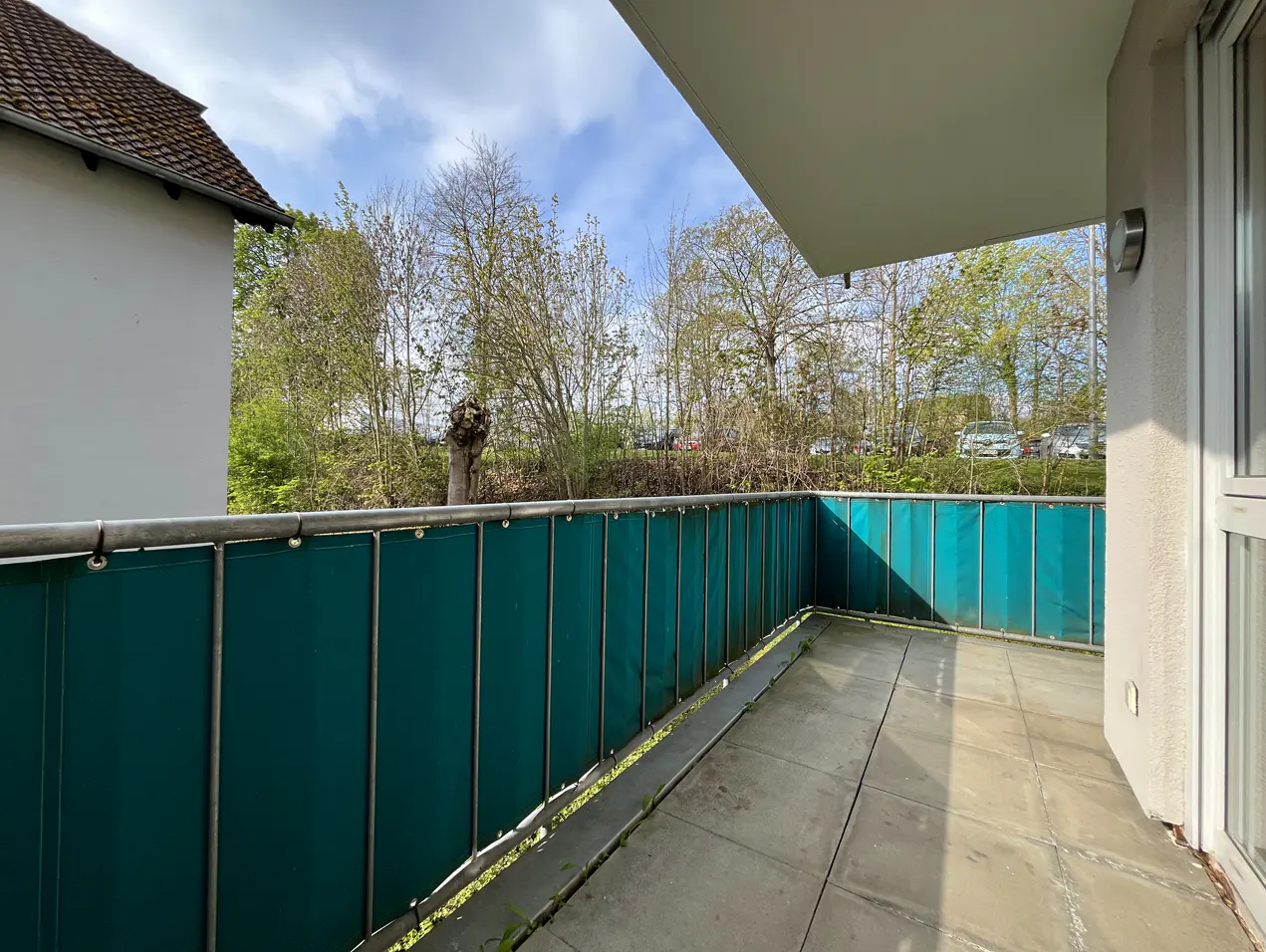 Balcony view with green privacy screen, metal railing, and gray tiled floor. Trees and a building are visible in the background.