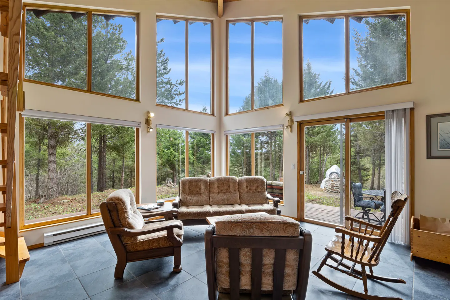 Living room with large windows showing trees, blue sky, brown sofa, chairs, and a rocking chair on a dark tile floor.