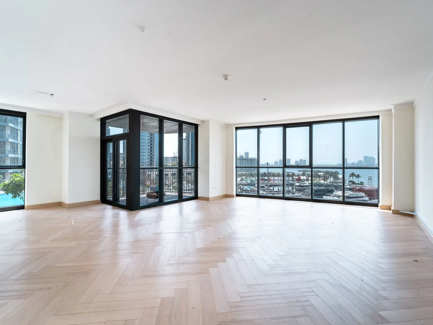 Bright, empty room with white walls, herringbone wood floors, and large windows showing a city and harbor view.