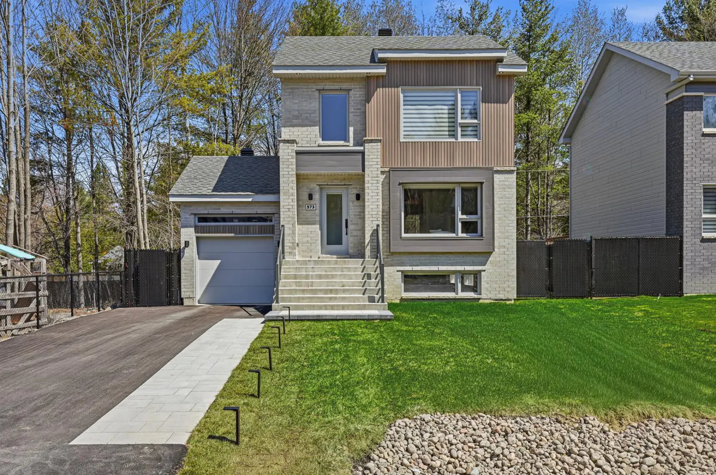 Two-story house with a gray brick and brown wood facade, a gray roof, and a white front door. A driveway and green lawn are in front.
