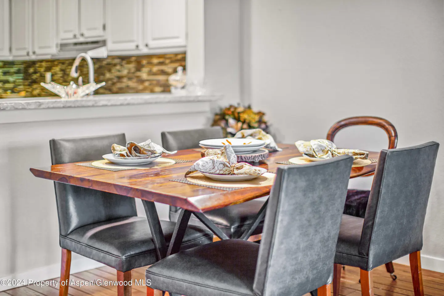 A dining room features a wooden table set for six with gray upholstered chairs. A kitchen is visible in the background.