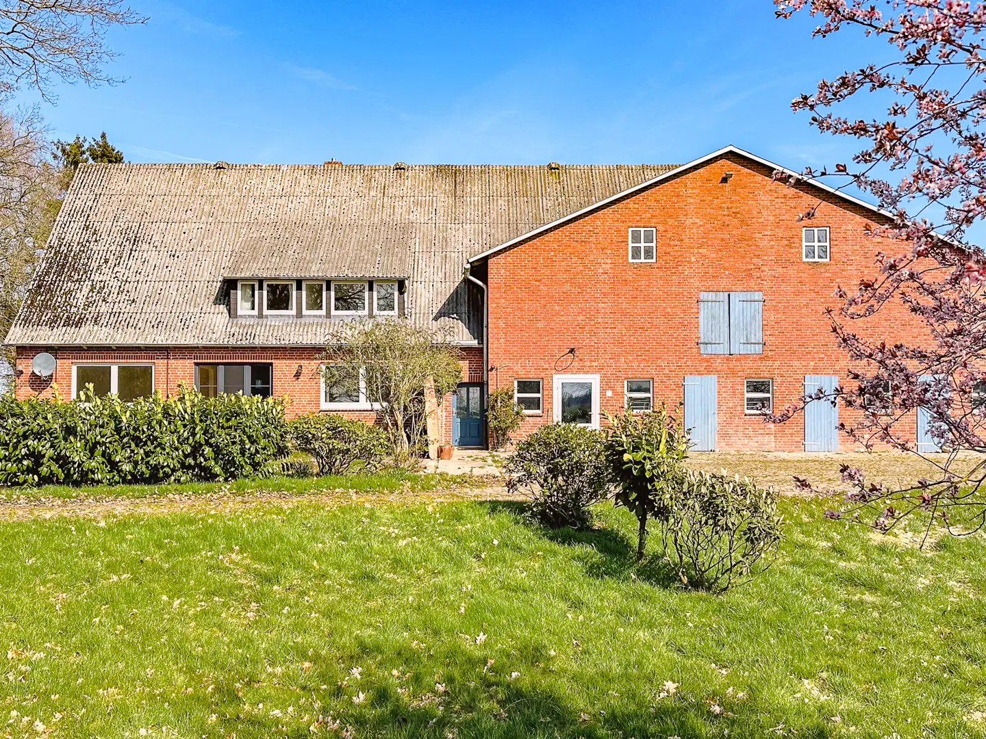 Exterior view of a red brick farmhouse with a gray roof, blue shutters, and a green lawn under a blue sky.