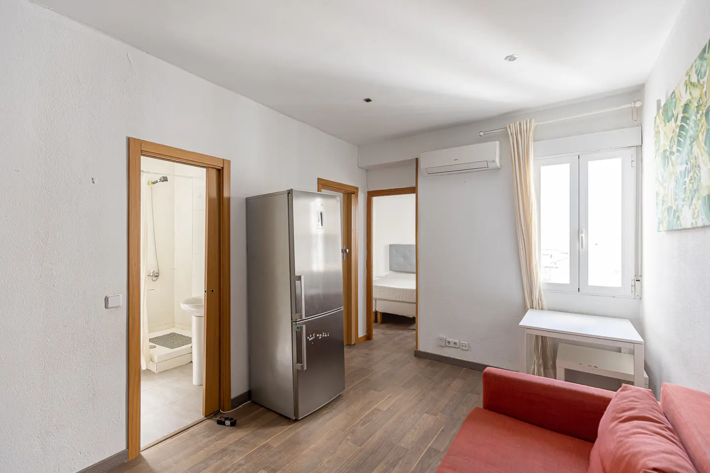 A bright apartment interior with a stainless steel refrigerator, a red sofa, and open doorways to a bathroom and bedroom.