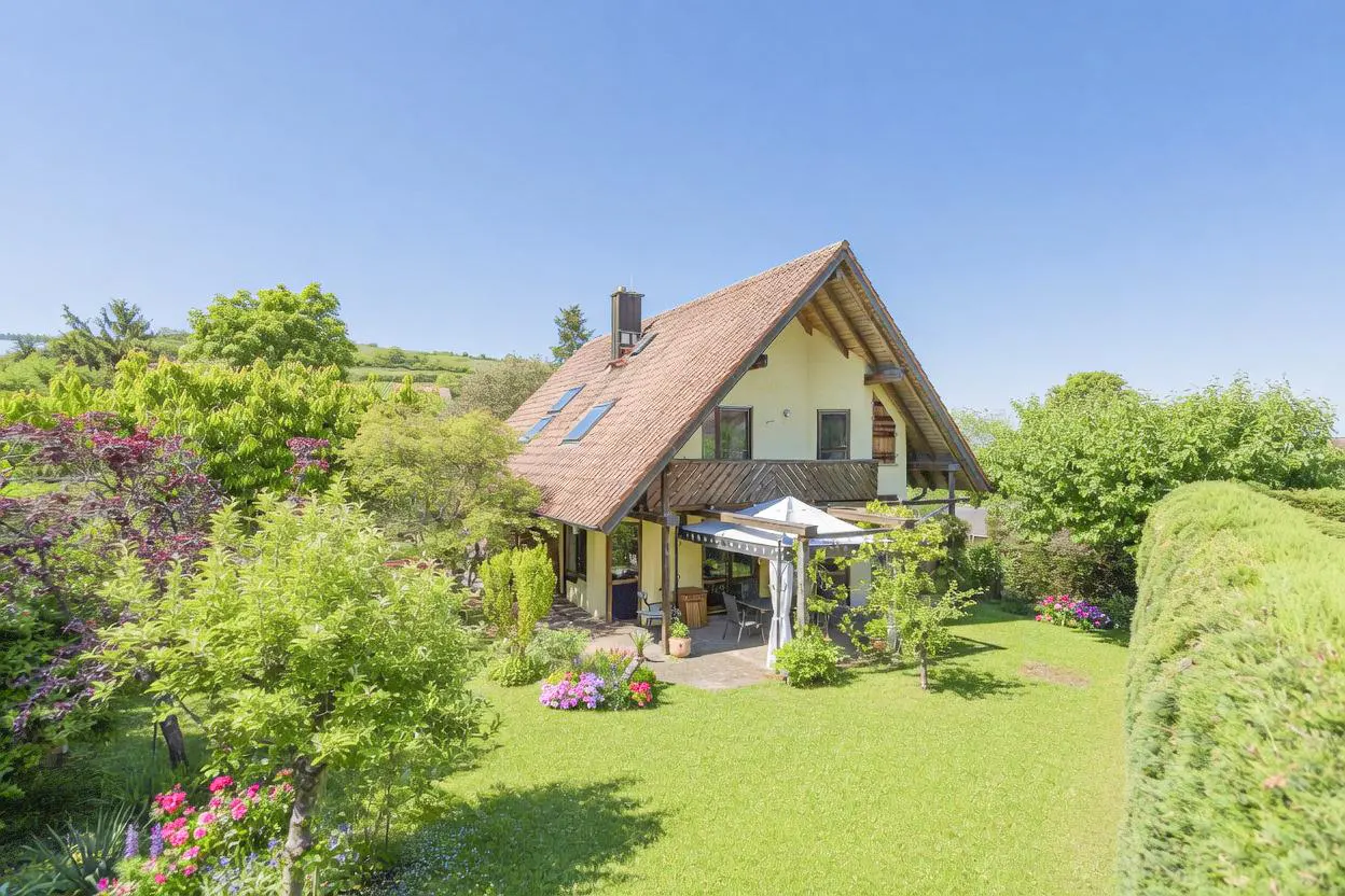 A charming yellow house with a red tile roof, surrounded by lush green trees and a well-manicured lawn under a clear blue sky.