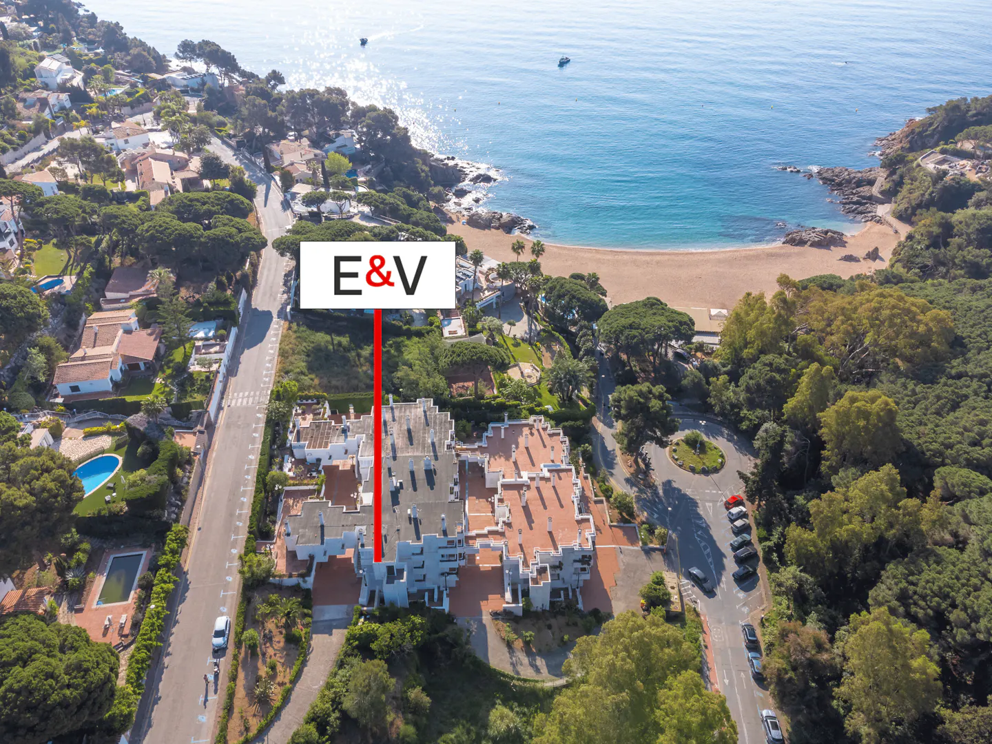 Aerial view of a multi-story building with a red tile roof, near a sandy beach and blue ocean. "E&V" logo is displayed above the building.