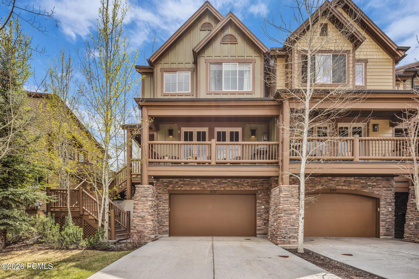 Two-story townhouses with stone accents, brown trim, and garages. Balconies with railings overlook the driveways. Blue sky background.