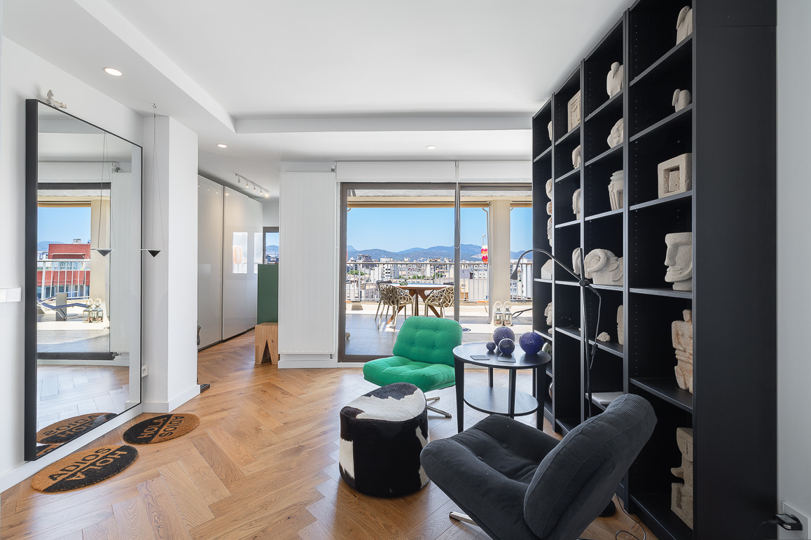 Modern living room with herringbone wood floors, a tall black bookshelf, green and grey chairs, and a balcony view. [AI-generated text]