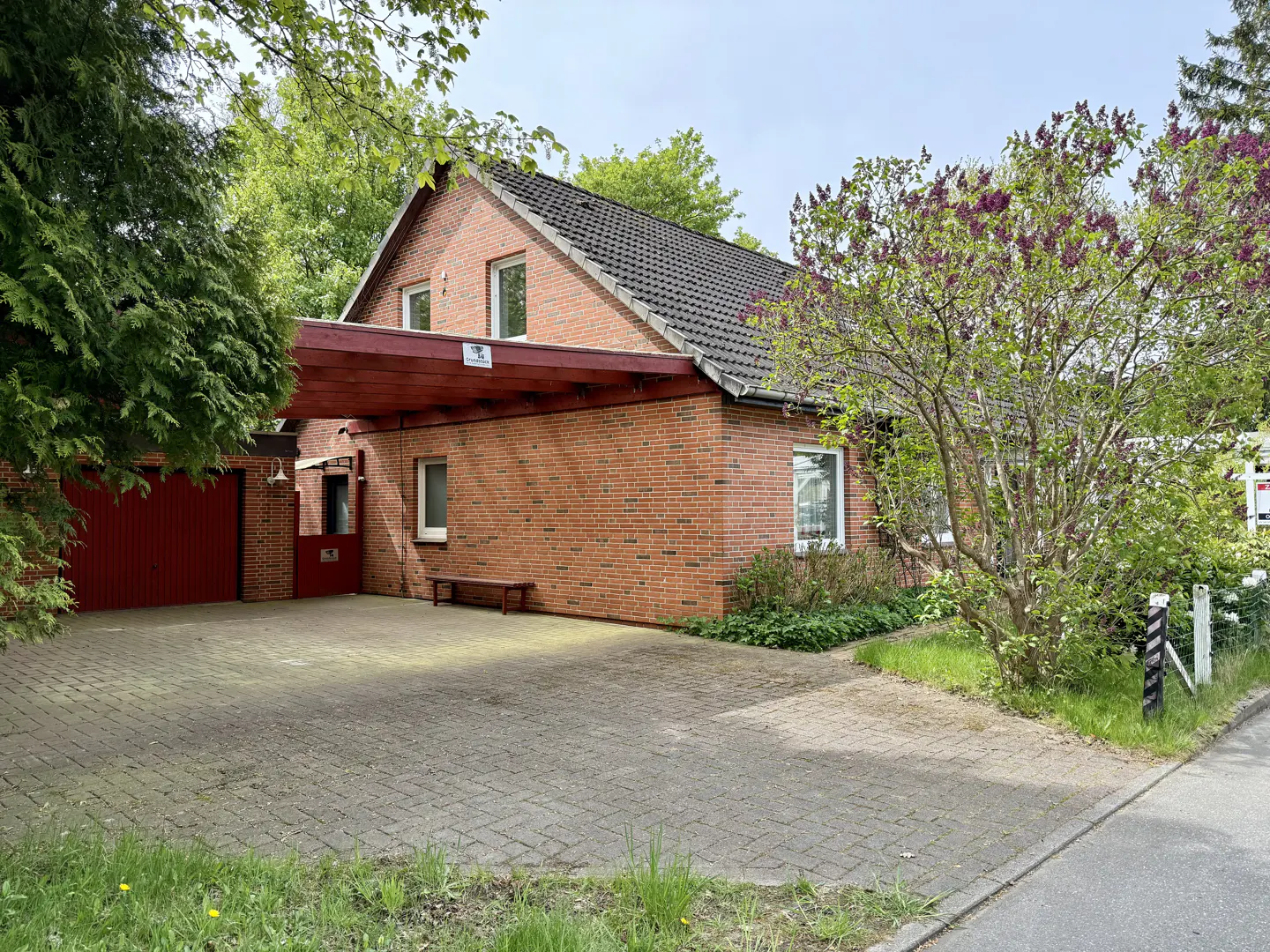 A single-story brick house with a dark roof and a red garage door is surrounded by green trees and a blooming lilac bush.