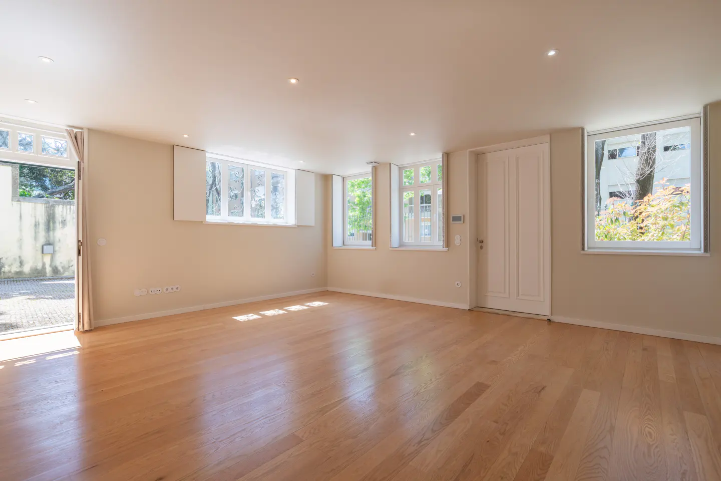 Bright, empty room with light wood floors, beige walls, and white trim. Windows and an open door let in natural light.