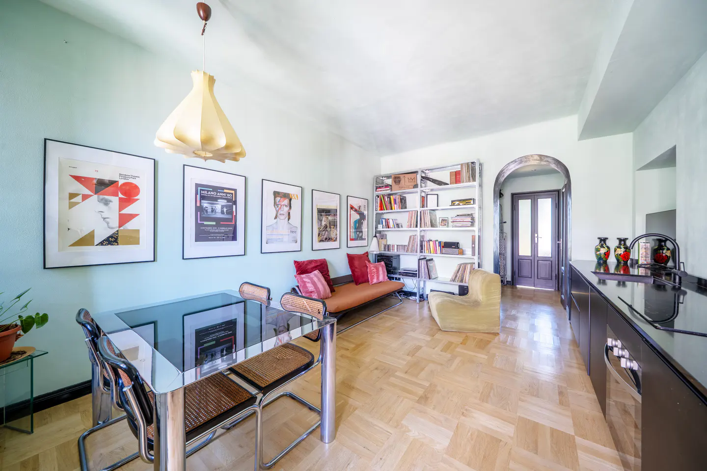Modern dining area with glass table, cane chairs, gallery wall of posters, orange bench, and open shelving on wood floors.