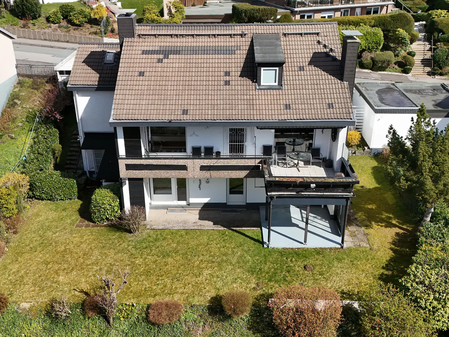Aerial view of a two-story white house with a brown tiled roof, chimney, and a covered patio, surrounded by a green lawn and trees.