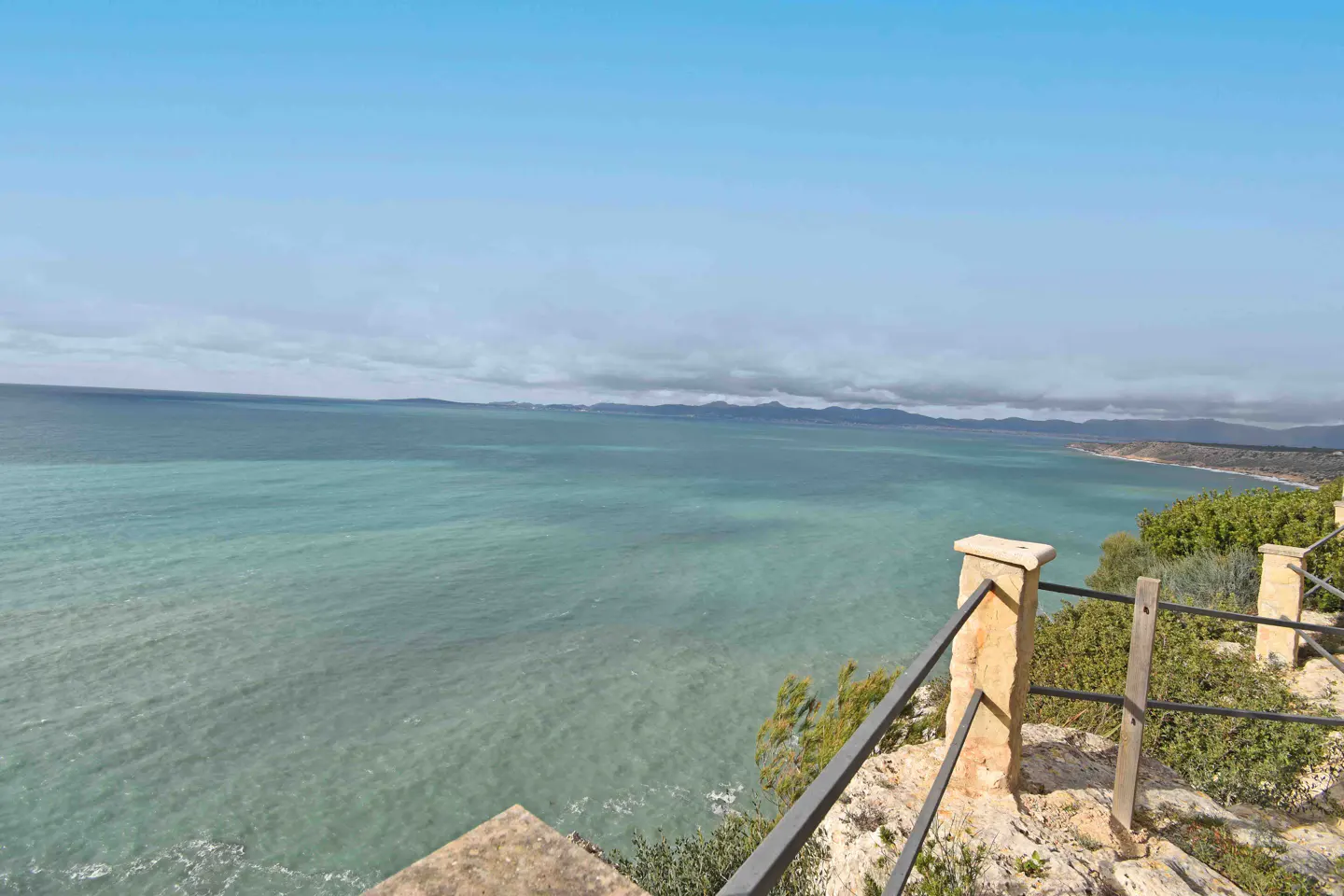 Ocean view from a stone terrace with a metal railing. Turquoise water stretches to the horizon under a blue sky.