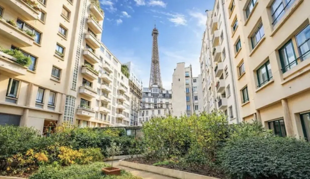 Paris apartment courtyard view with the Eiffel Tower in the background. Beige buildings surround green bushes under a blue sky.