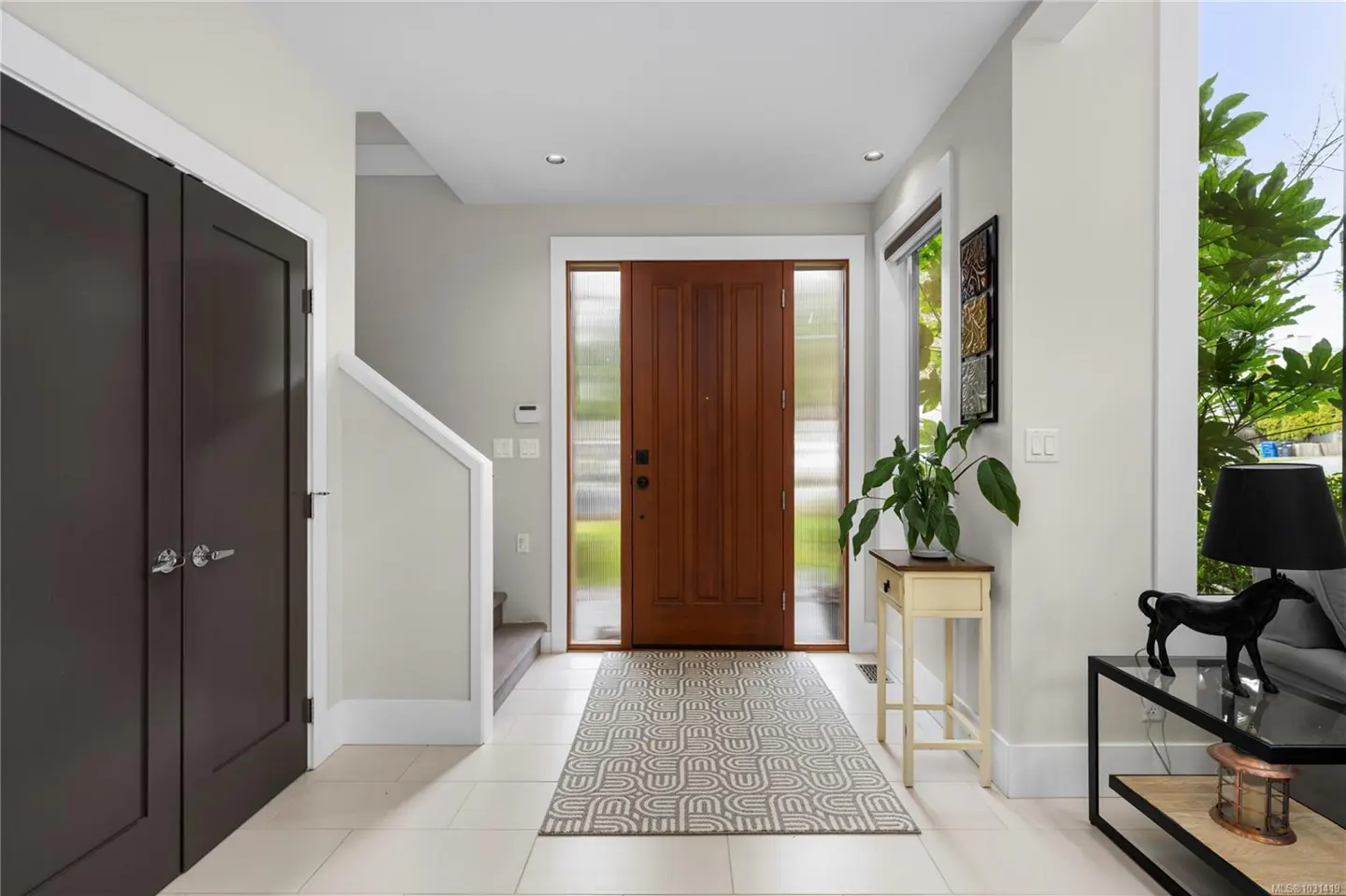 A bright foyer with a wood front door, a patterned rug, and a black horse statue on a glass table.