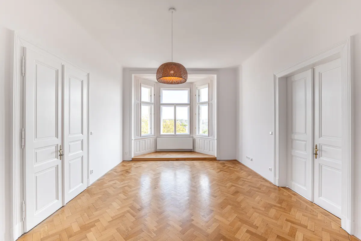 Bright, empty room with herringbone wood floors, white walls, and a bay window. A woven pendant light hangs from the ceiling.